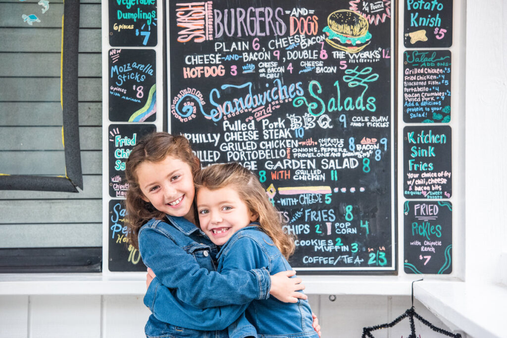 Two small girls hugging in front of a colorful chalkboard menu 
