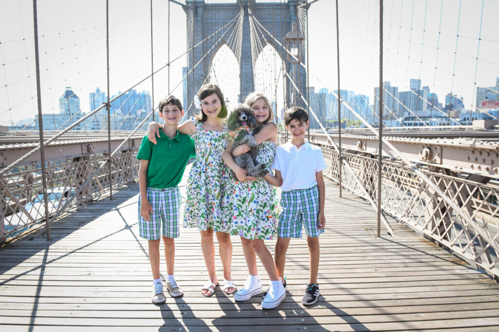 Four kids smiling on the Brooklyn Bridge while one holds a dog 