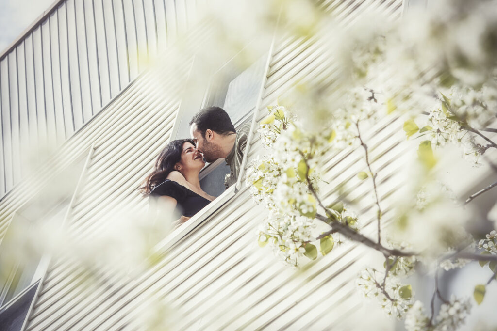 A couple leans out of a window of a building, sharing a kiss surrounded by blooming branches.