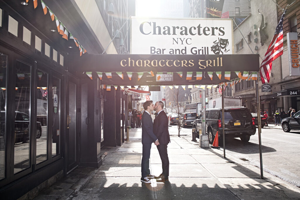 A couple in suits stand face-to-face, kissing, under the sign for "Characters NYC Bar and Grill," with city street activity in the background.