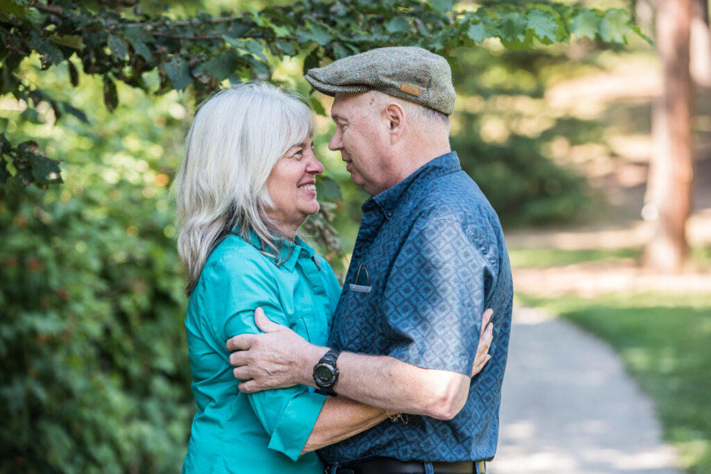 A couple stands facing each other, holding hands and smiling, in a green, leafy outdoor setting.