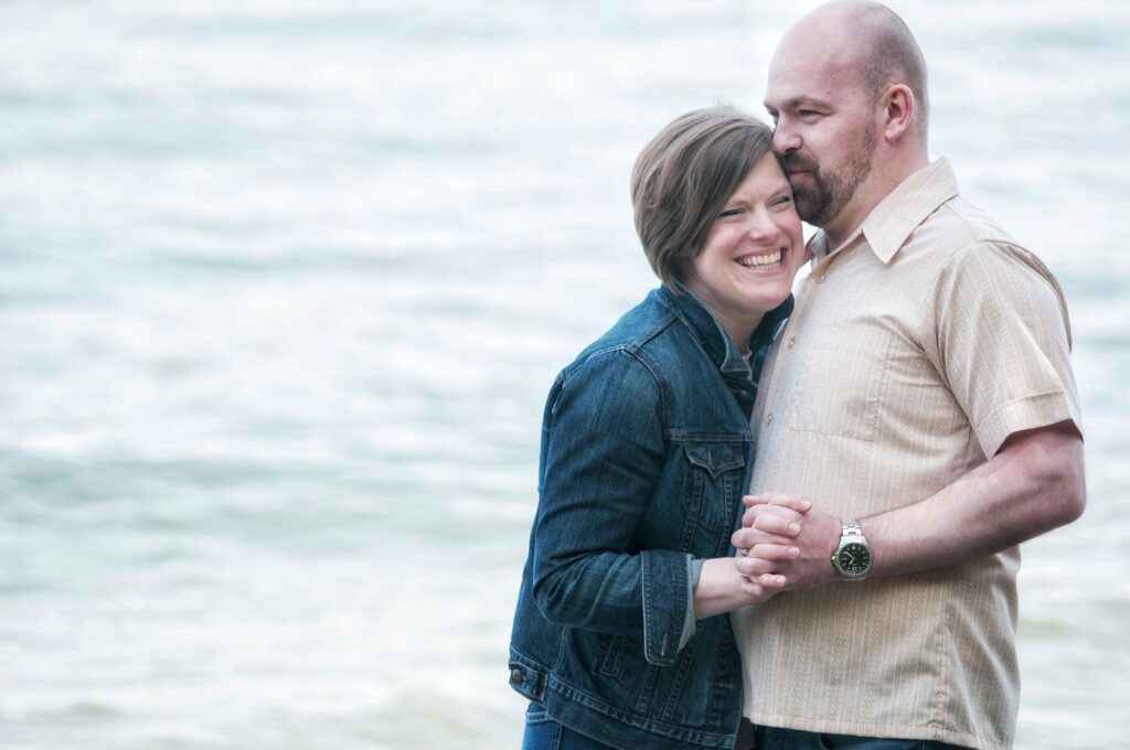 A couple stands close together by the water, smiling and holding hands, with the man gently kissing the woman's forehead.