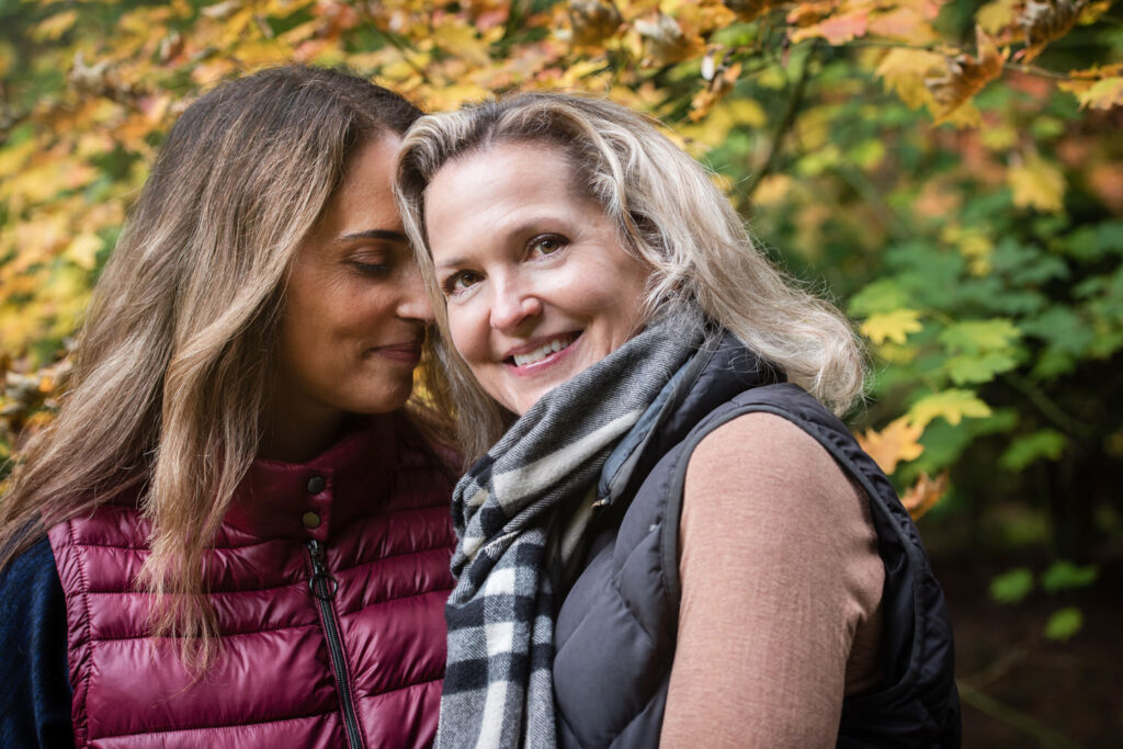 A couple with two women stand closely together, one with her head resting on the other's shoulder, smiling amidst a backdrop of autumn foliage.