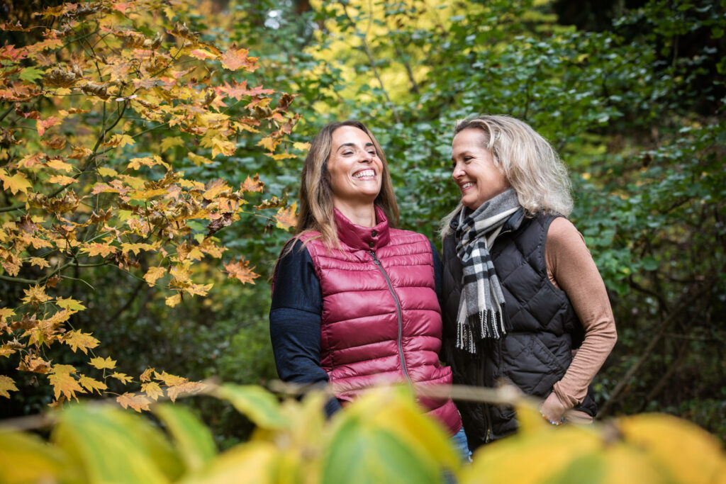 A couple laughs together, surrounded by vibrant autumn leaves, both dressed warmly in puffy vests and scarves.