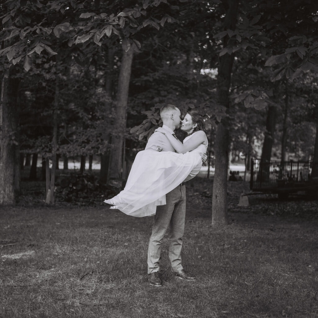 A man in dress pants and a shirt lifts and kisses a woman in a white dress in a wooded area, with trees in the background.