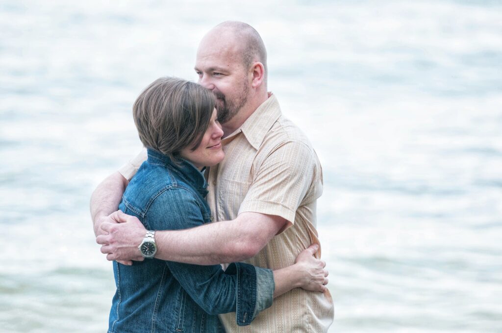 A couple embraces by the water, with one person wearing a denim jacket and the other in a light shirt, both appearing content and affectionate.