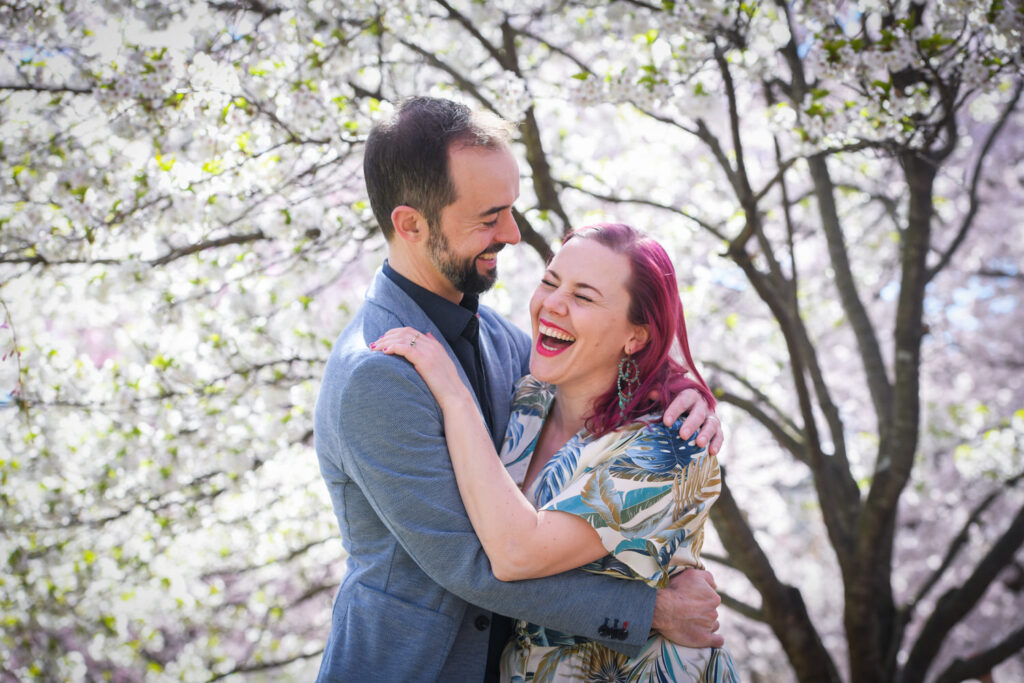 A couple embraces and laughs under blooming trees, with the woman wearing a patterned dress and the man in a suit jacket.