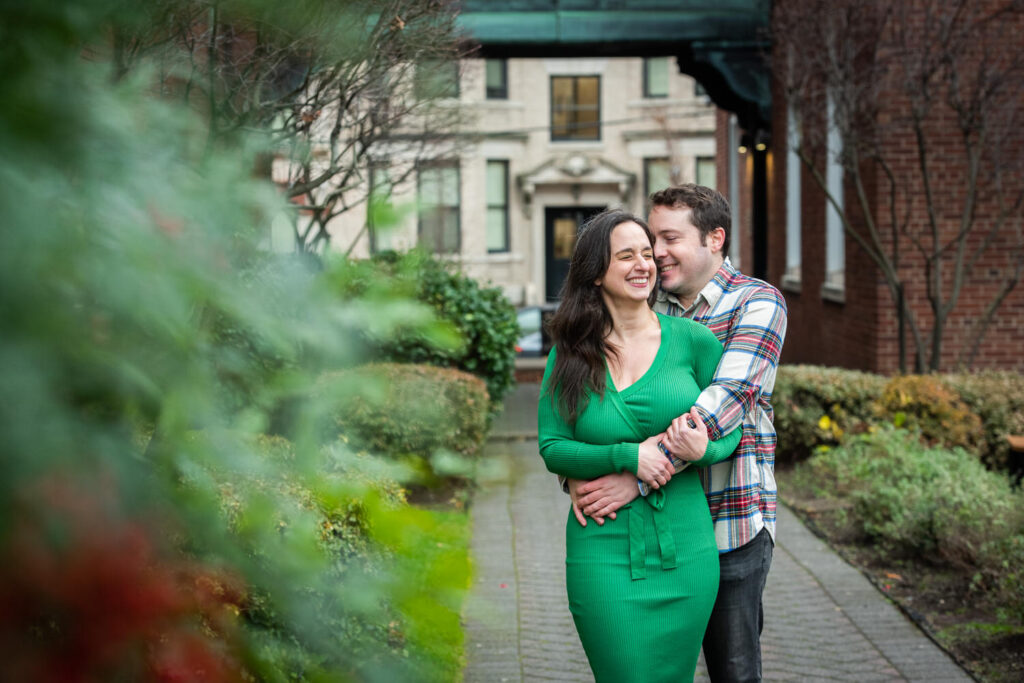 A man in a plaid shirt hugs a woman in a green dress from behind, both smiling and standing on a garden path with buildings in the background.