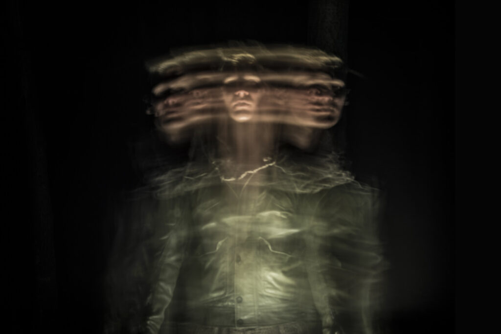 A person appears blurred in motion, with multiple overlapping faces, creating a surreal and mysterious effect in a dark setting.