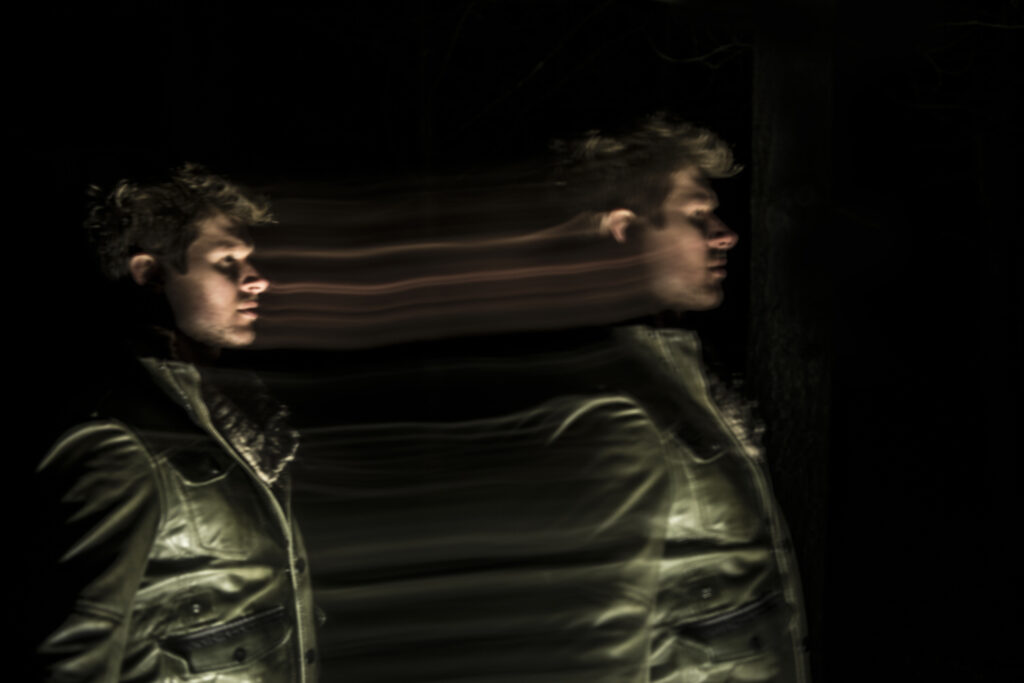 A man's profile is duplicated with a motion blur effect, giving the appearance of two faces in a dark setting.