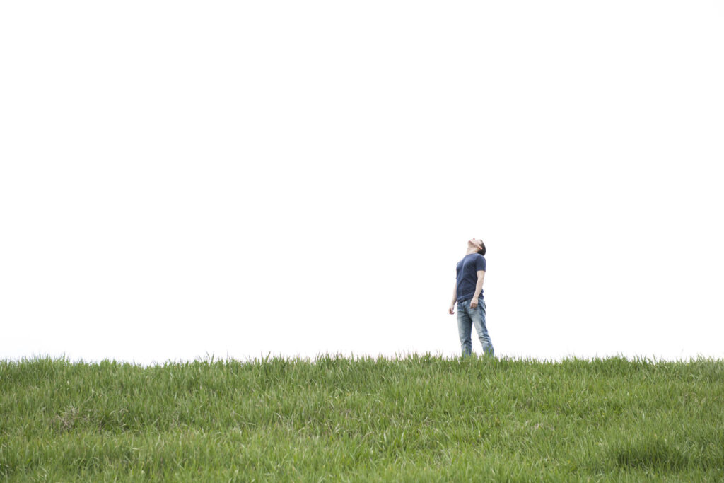 A person stands on a grassy hill, looking up at the sky with a serene expression, against a backdrop of an overcast white sky.