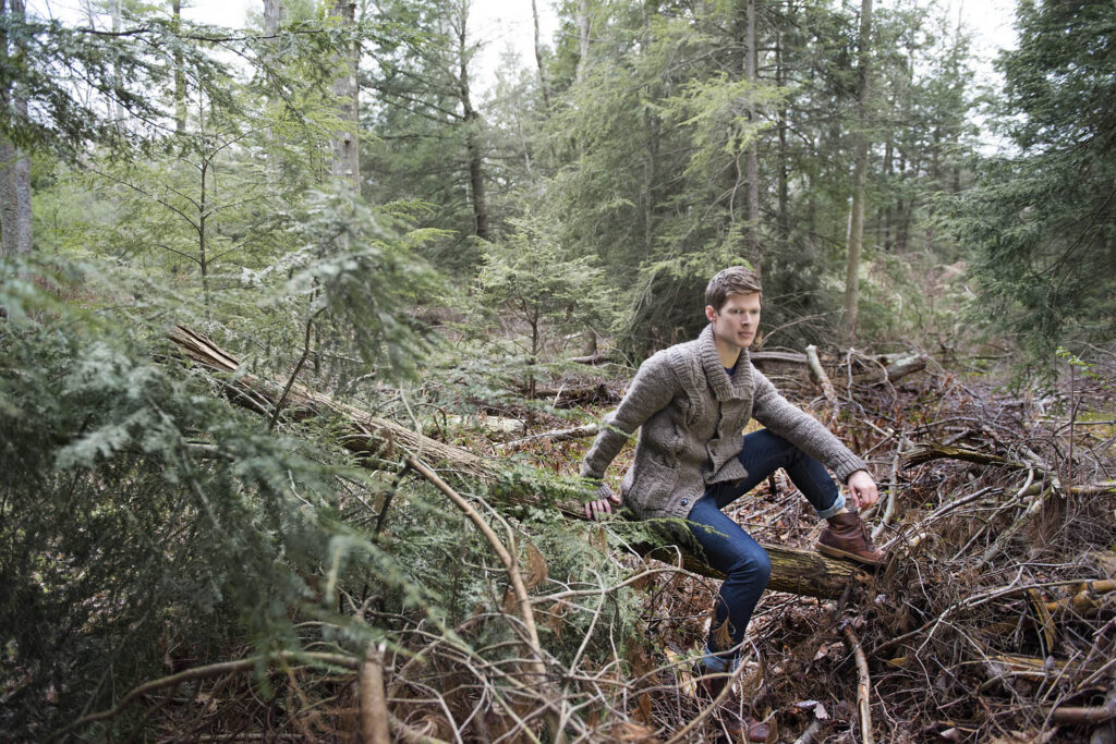 A person sits on a fallen tree in a dense forest, wearing a grey sweater and jeans, looking thoughtfully into the distance.