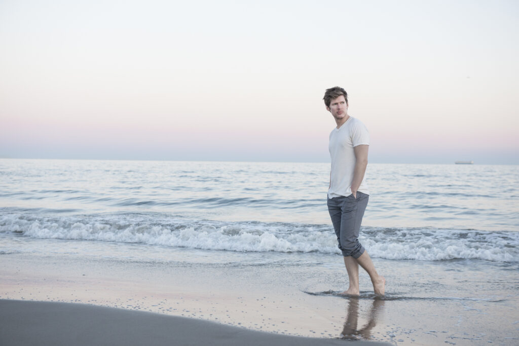 A man walks barefoot along the shore, with gentle waves and a pastel sunset in the background, wearing rolled-up pants and a white t-shirt.