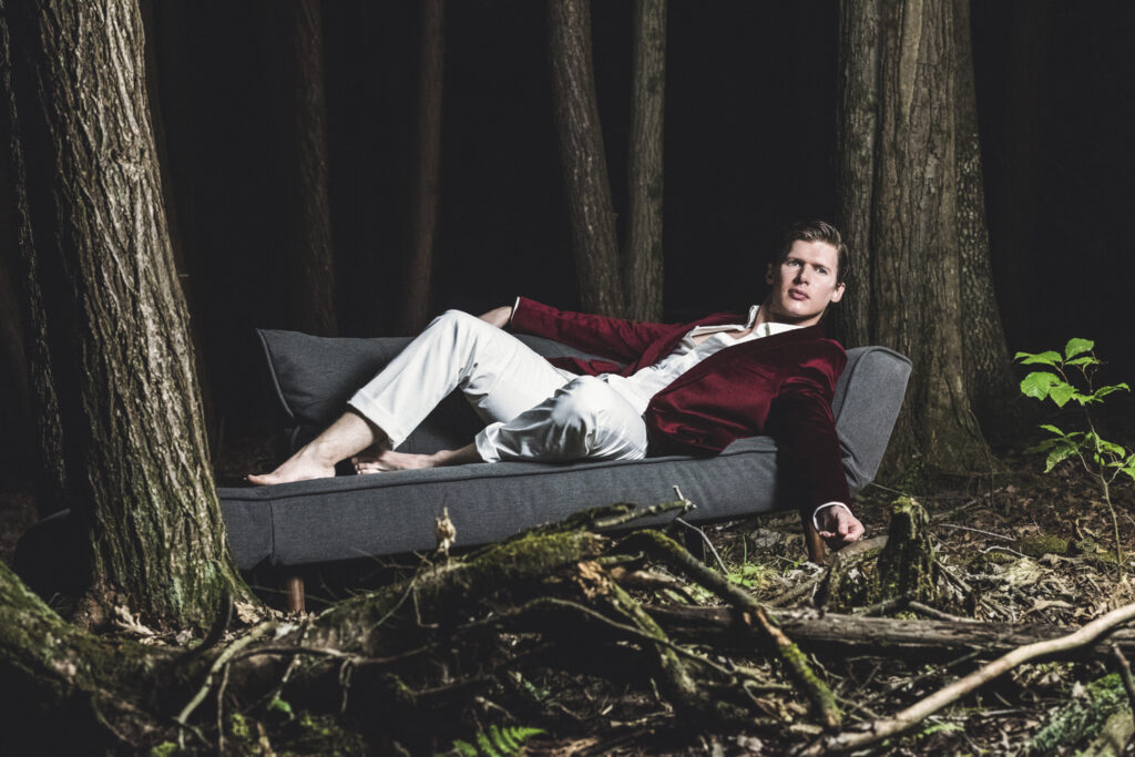 A man in a burgundy blazer and white pants lounges on a grey couch in a forest at night.