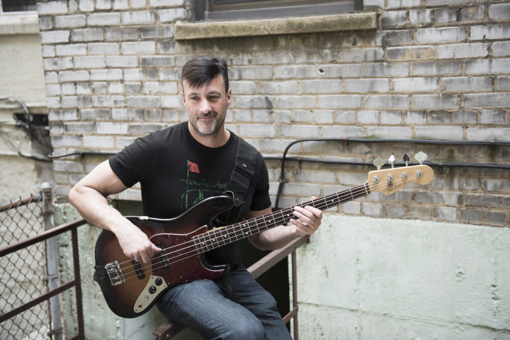 A man sits with a bass guitar in an outdoor setting against a backdrop of weathered brick walls.