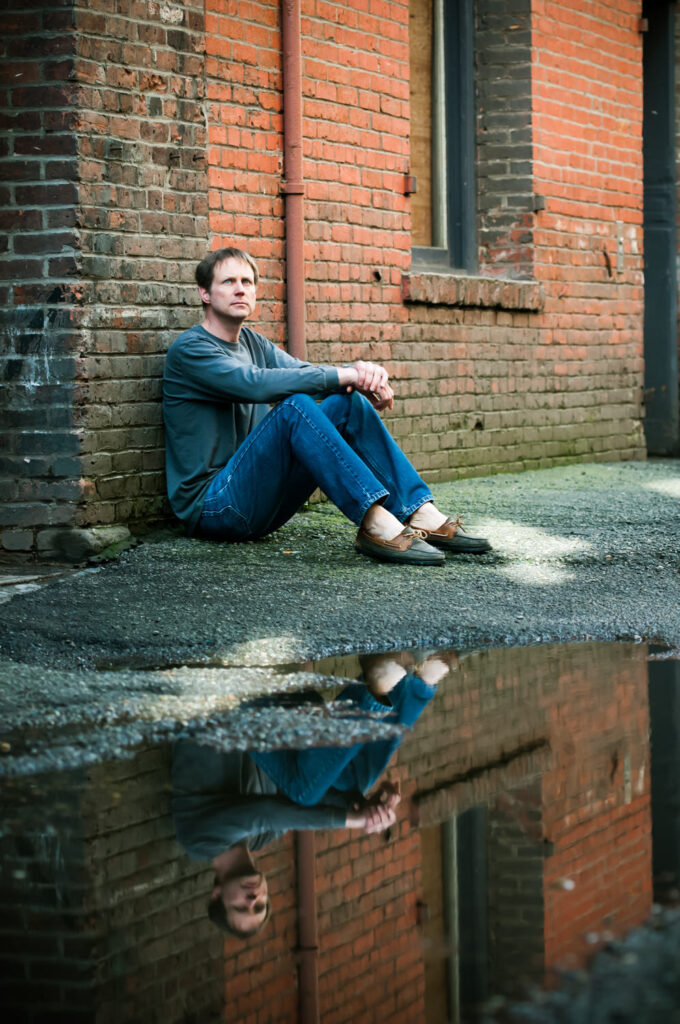 A man sits against a brick wall, reflected in a nearby puddle, looking contemplative.