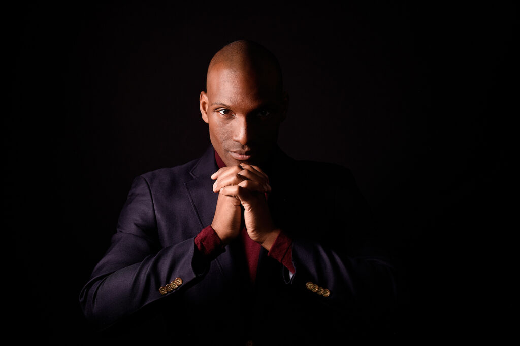 A man in a dark suit and maroon shirt poses with hands clasped under his chin, set against a dark background.