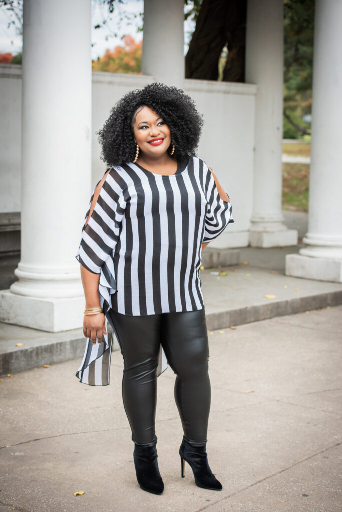 A woman with curly hair, dressed in a black and white striped top and black leggings, poses confidently outdoors in front of white columns.