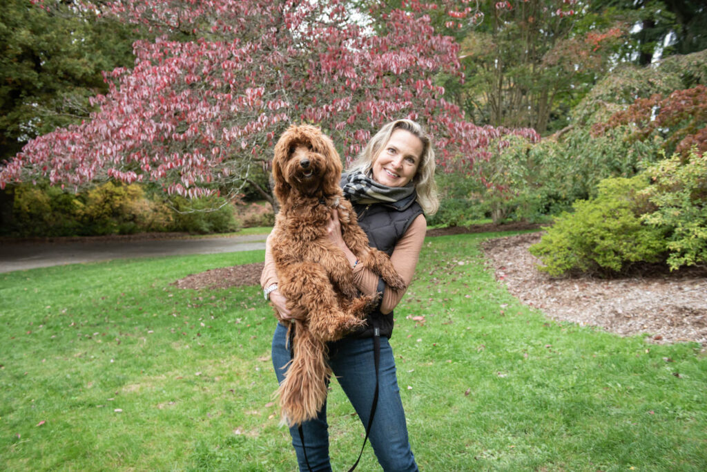 A woman stands in a park holding a fluffy brown dog, with trees featuring autumn foliage in the background.