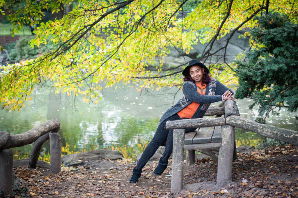 A woman in an orange top and black hat leans on a rustic wooden bench by a pond, surrounded by vibrant autumn foliage.