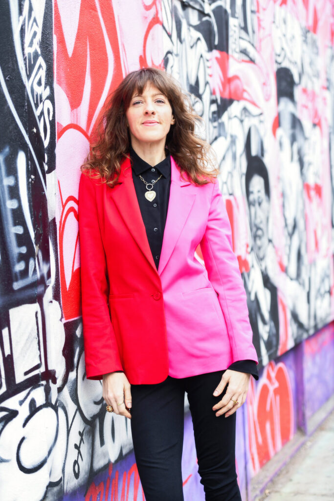 A woman in a vibrant red and pink jacket leans against a graffiti-covered wall, exuding confidence.