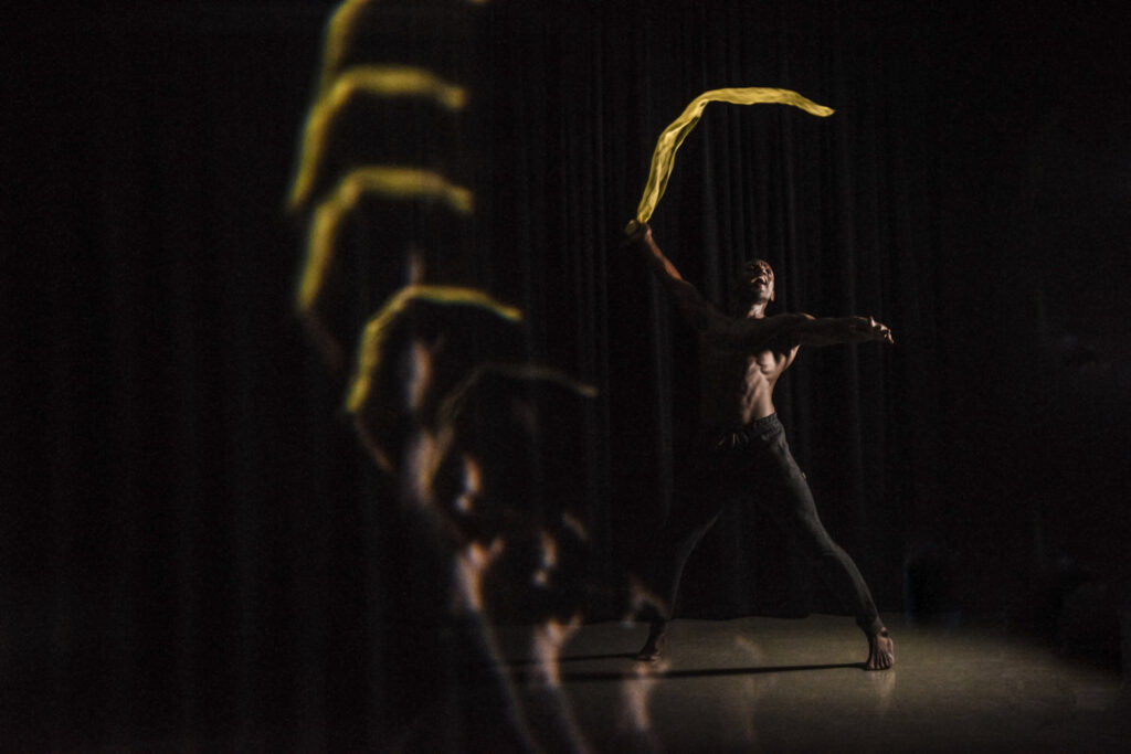 A man performs a dynamic dance move with a yellow ribbon, creating a motion blur effect in a dark setting.