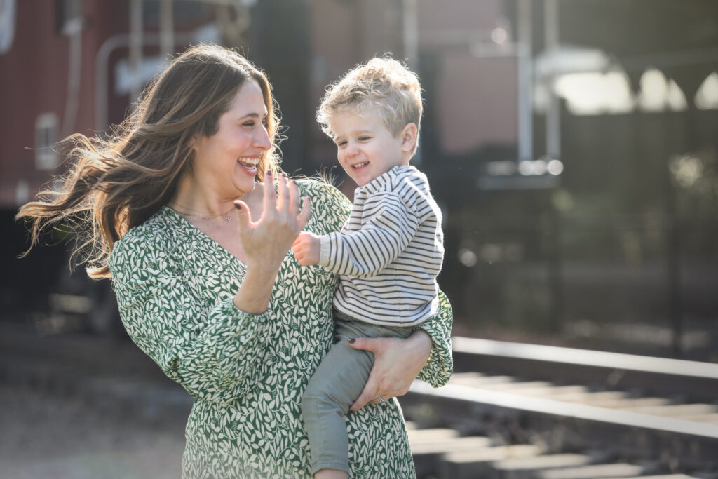 A mother in a green patterned dress joyfully interacts with her toddler, who is wearing a striped shirt, in front of an outdoor train station.