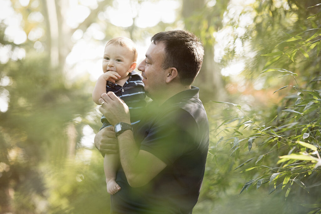 A father holds his baby in a lush, green outdoor setting during golden hour. The baby is wearing a striped shirt and has his hand near his mouth.