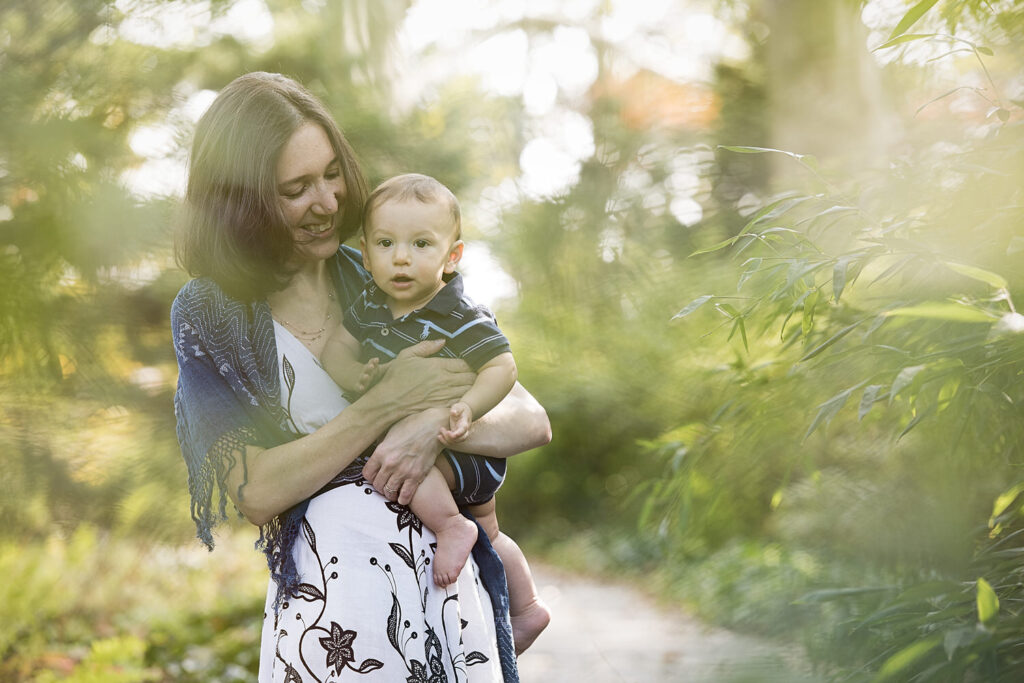 A mother cradles her baby while standing on a garden path surrounded by greenery. 