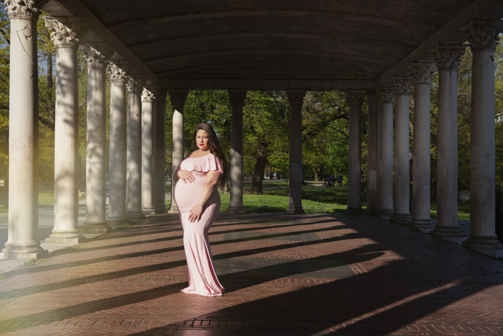 A pregnant woman in a flowing pink dress stands under a columned archway in a park, bathed in the soft light of golden hour.