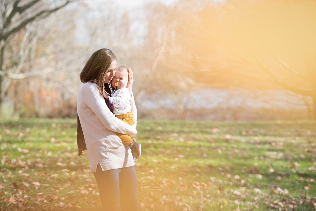 A mother holds her baby in an autumn park, with the baby wrapped in warm clothes and snuggled close as they enjoy the golden light.