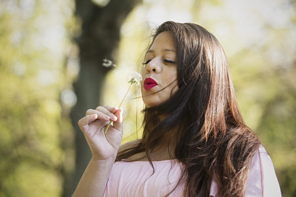 A woman with long hair blows on a dandelion, sending its seeds into the air. 