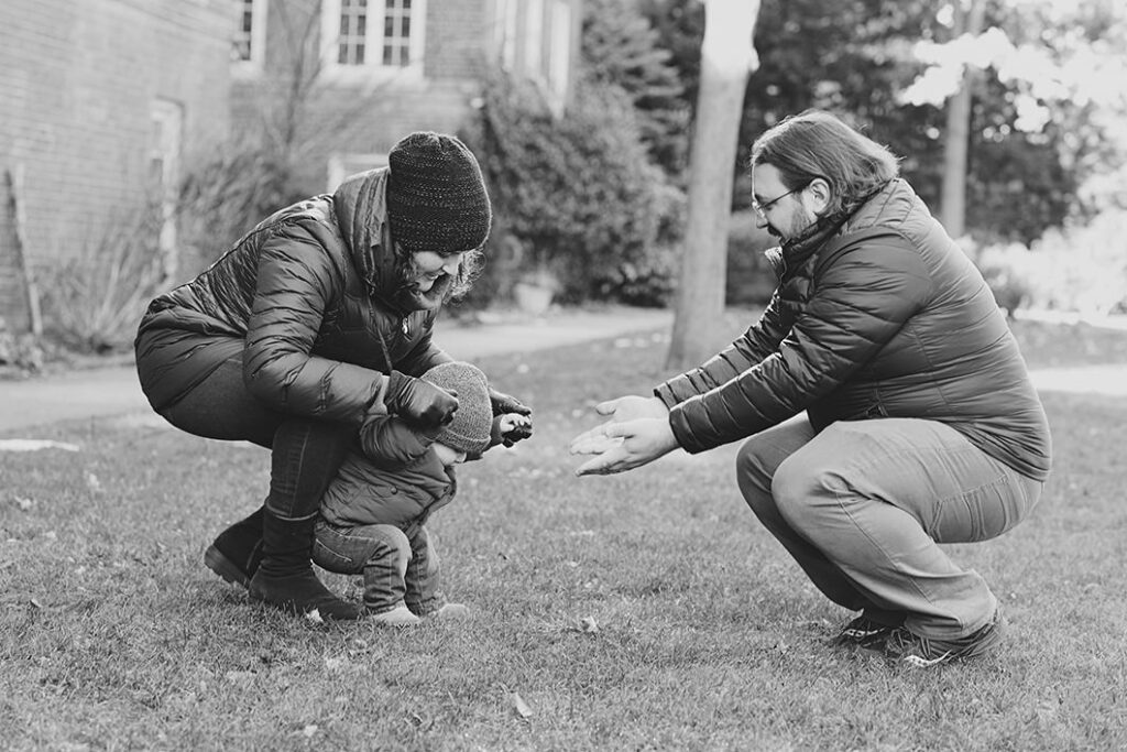 A parent reaching out towards their baby as the other parent helps the baby stand up 